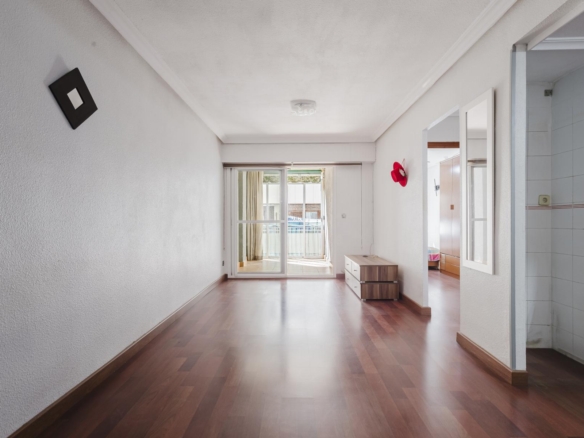 Bright empty living room with sliding doors to balcony, wooden floors, and a small dresser along the right wall; red hat mounted on wall.