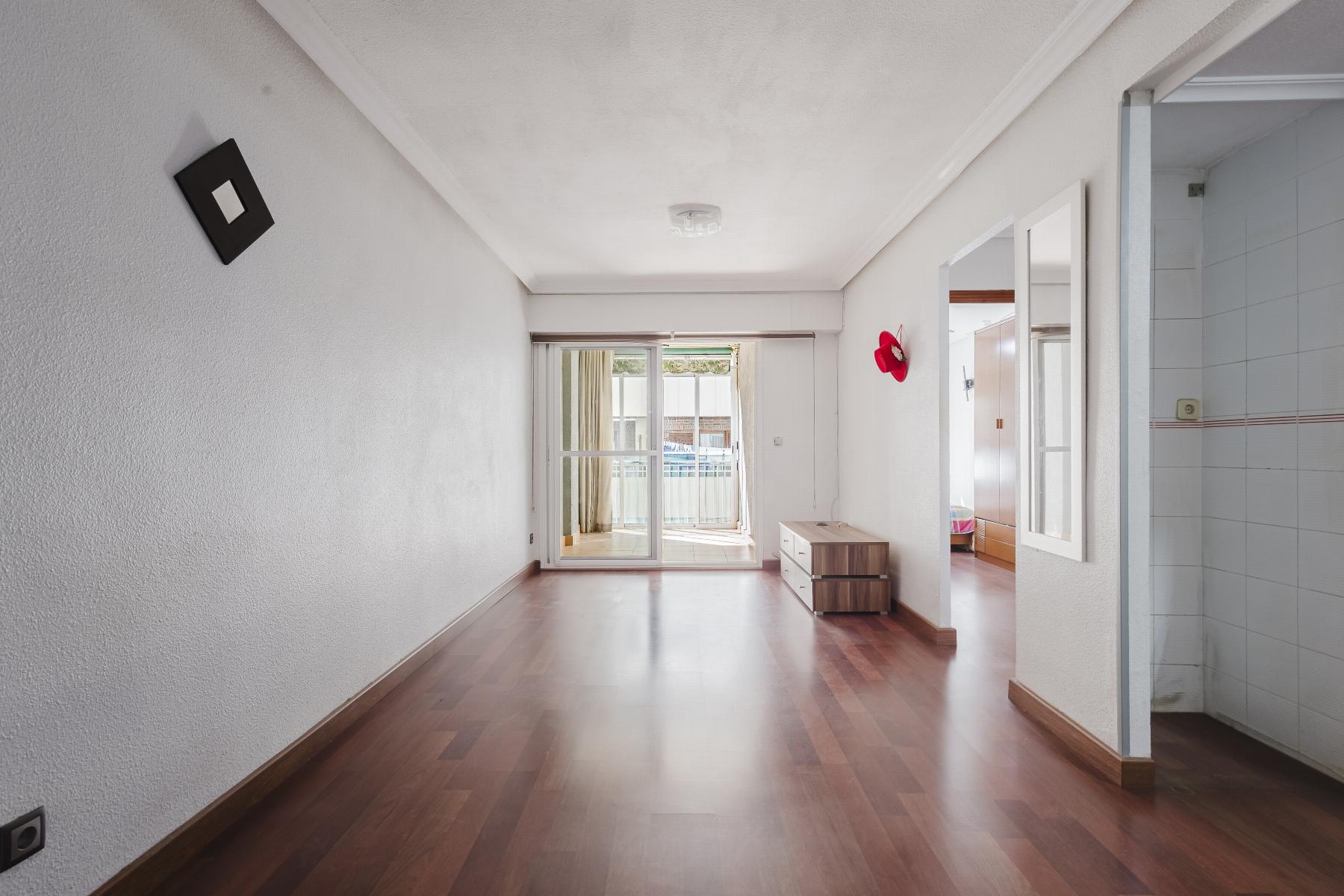 Bright empty living room with sliding doors to balcony, wooden floors, and a small dresser along the right wall; red hat mounted on wall.