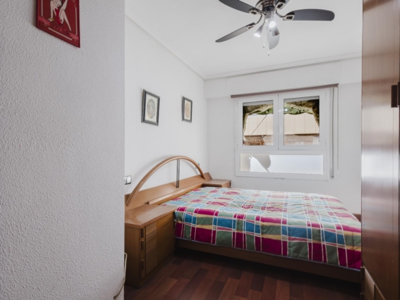 Small bedroom with a wooden bed, curved headboard, and a colorful plaid quilt, plus a nightstand and wall art. A ceiling fan and window are in view.
