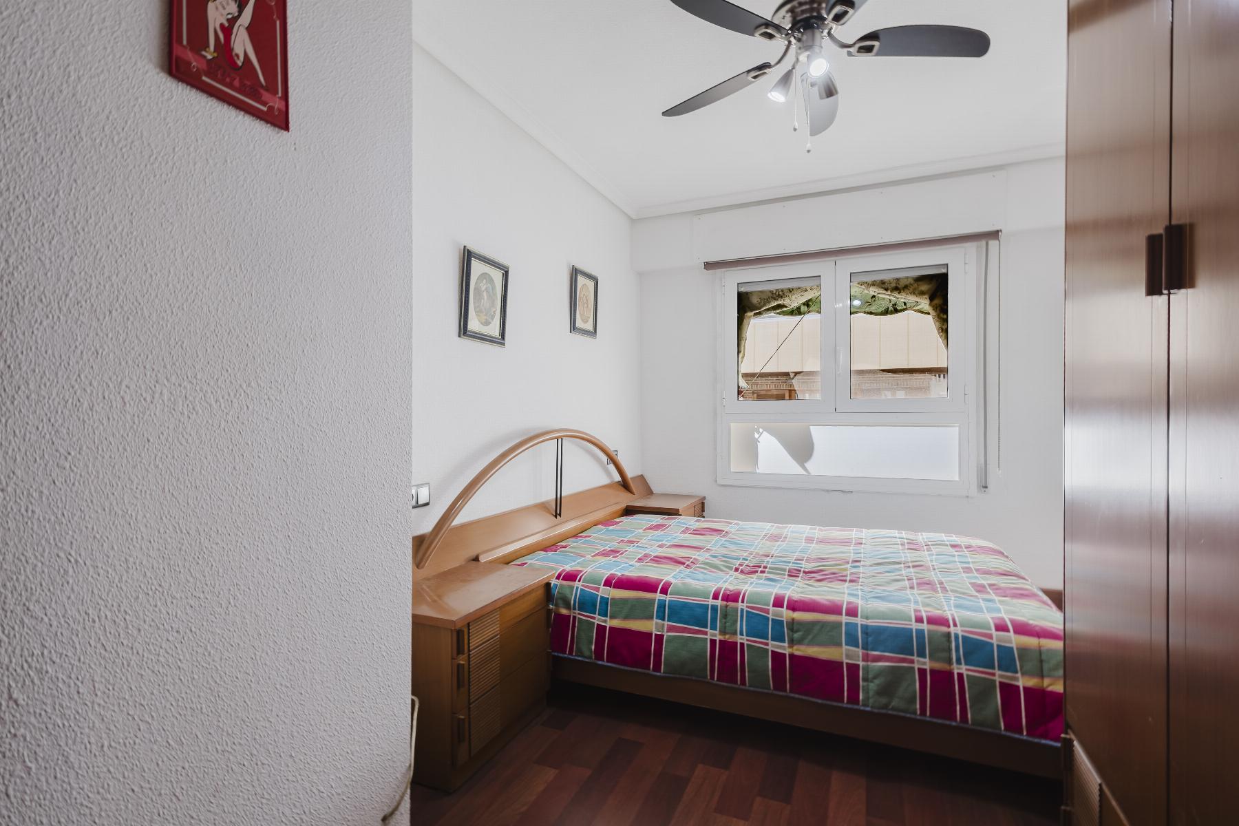 Small bedroom with a wooden bed, curved headboard, and a colorful plaid quilt, plus a nightstand and wall art. A ceiling fan and window are in view.
