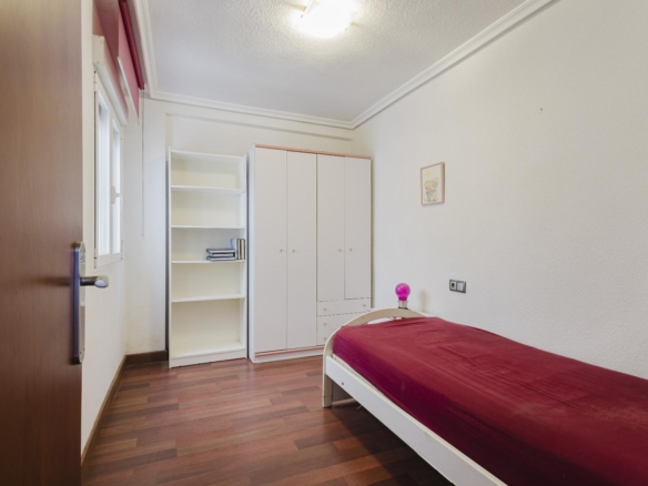 Small bedroom with white wardrobe and open bookshelf, a single bed with burgundy sheets, and a pink lamp on the bed. Wooden floor and white walls add brightness.