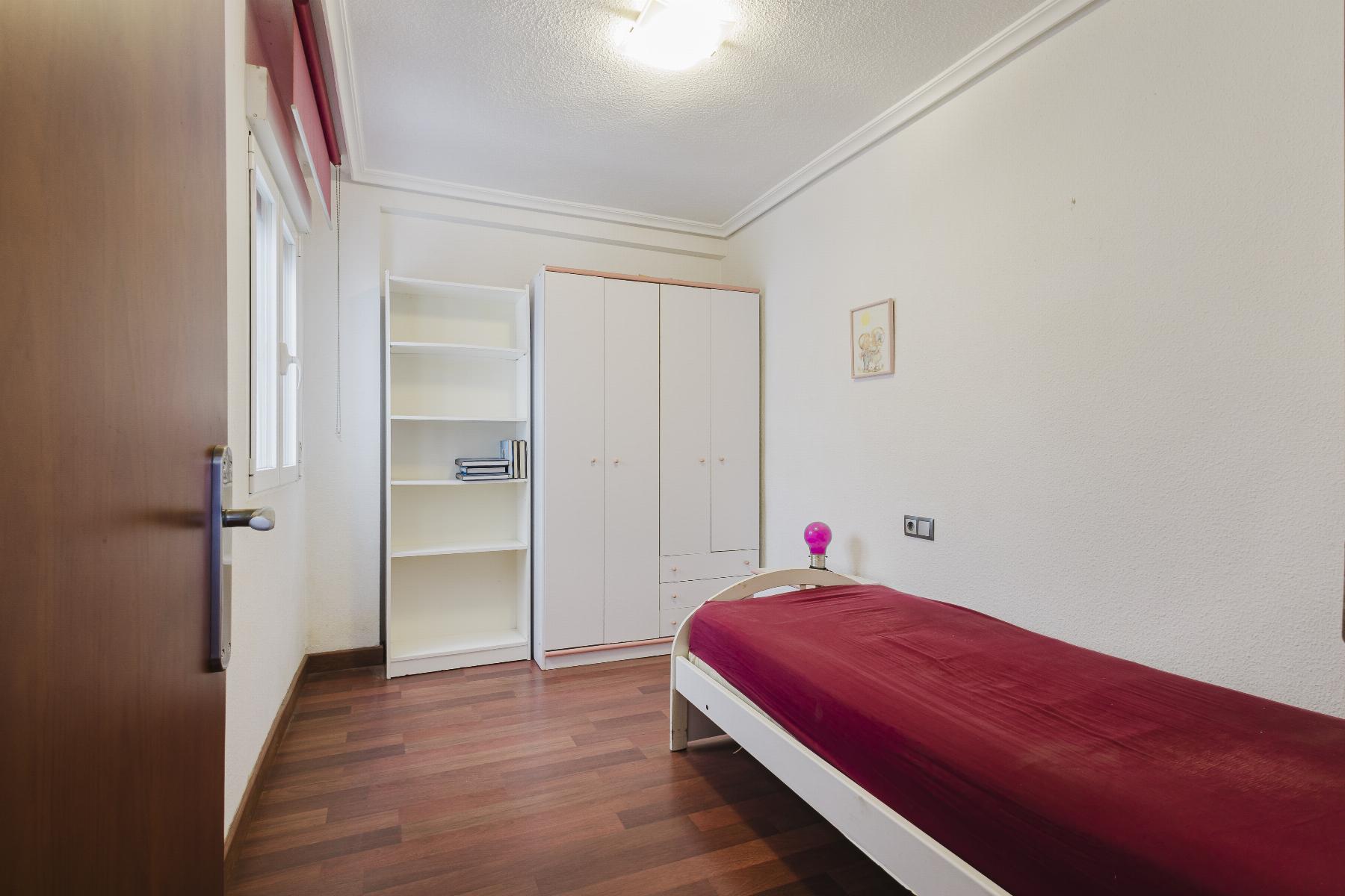 Small bedroom with white wardrobe and open bookshelf, a single bed with burgundy sheets, and a pink lamp on the bed. Wooden floor and white walls add brightness.