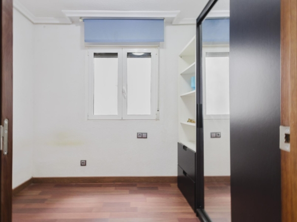 Small bright room with white walls, window with blue shade, and white shelving beside a dark sliding-door wardrobe; wooden floor.
