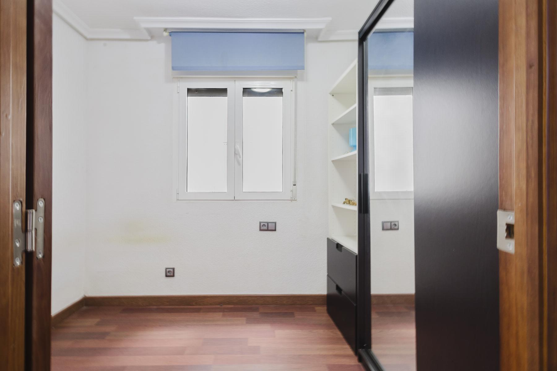 Small bright room with white walls, window with blue shade, and white shelving beside a dark sliding-door wardrobe; wooden floor.