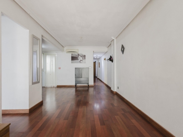 Long empty hallway in a home with white walls and dark wooden floor; a metal cabinet and a black-and-white photo are at the far end.