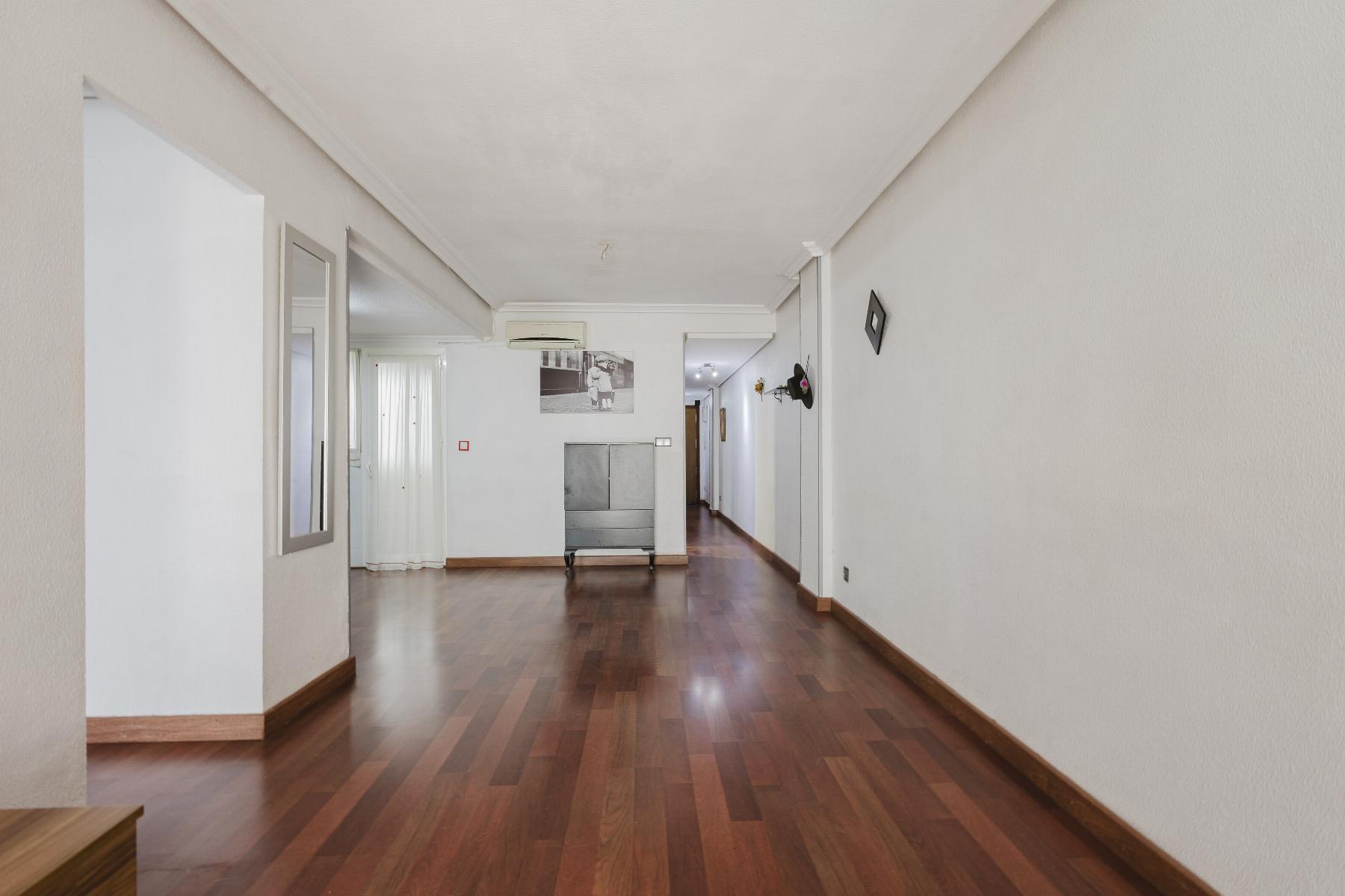 Long empty hallway in a home with white walls and dark wooden floor; a metal cabinet and a black-and-white photo are at the far end.