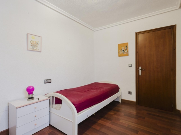 Small bedroom with white bed and dresser, burgundy duvet, pink lamp, and wall art; wooden door on the right.