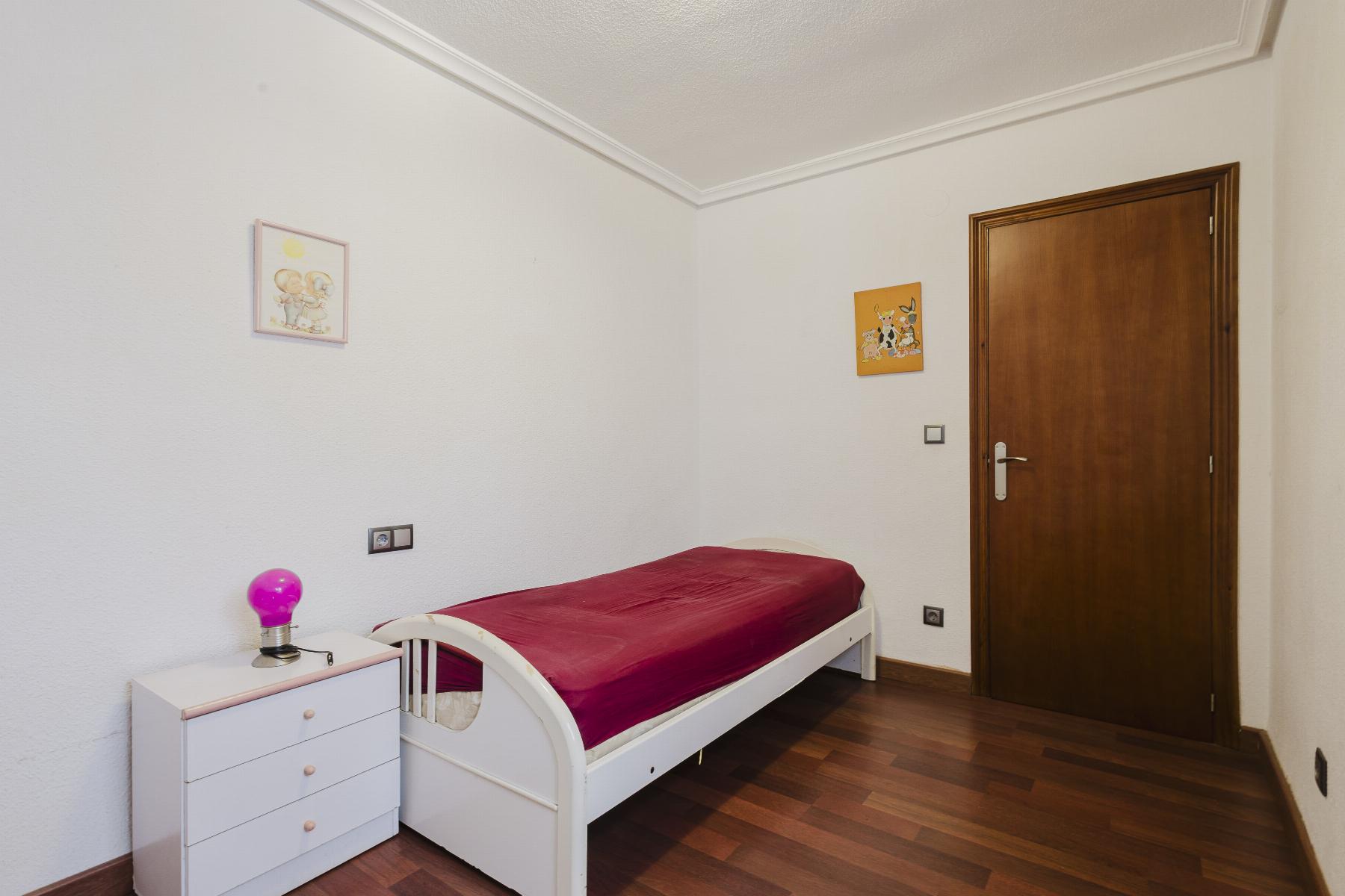 Small bedroom with white bed and dresser, burgundy duvet, pink lamp, and wall art; wooden door on the right.