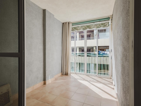 Sunlit balcony with beige tiled floor, gray walls, and large sliding glass doors opening to an outdoor pool area.