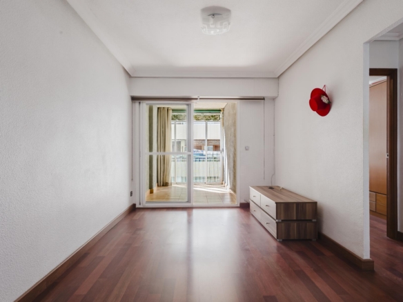 Empty living room with wood flooring, white walls, and a red hat hanging on the right wall near a dresser; sliding glass doors lead to a balcony.