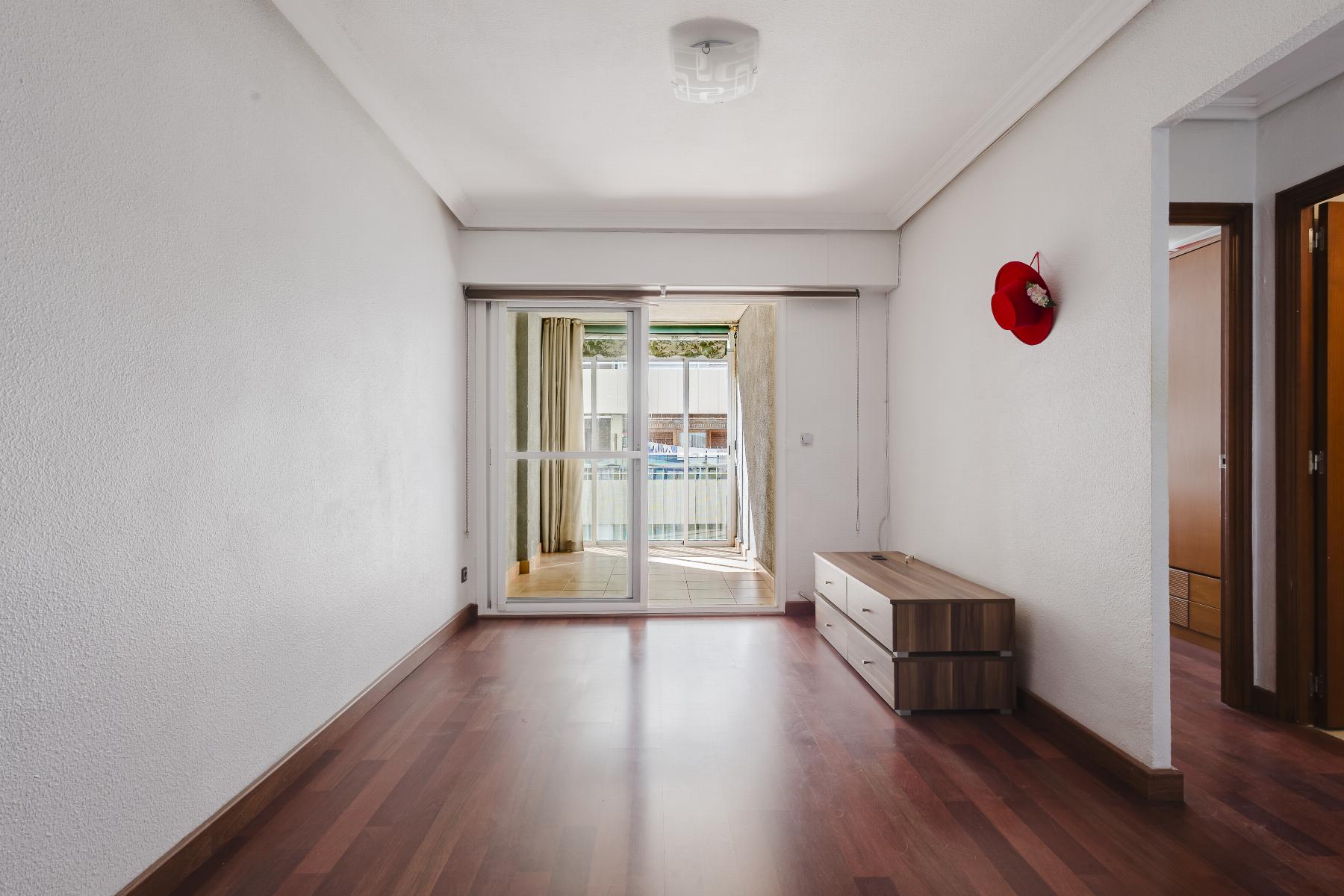 Empty living room with wood flooring, white walls, and a red hat hanging on the right wall near a dresser; sliding glass doors lead to a balcony.