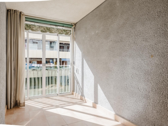 Sunlit room with beige curtains beside a sliding glass door opening to a balcony; textured gray walls and tiled floor casting striped shadows.