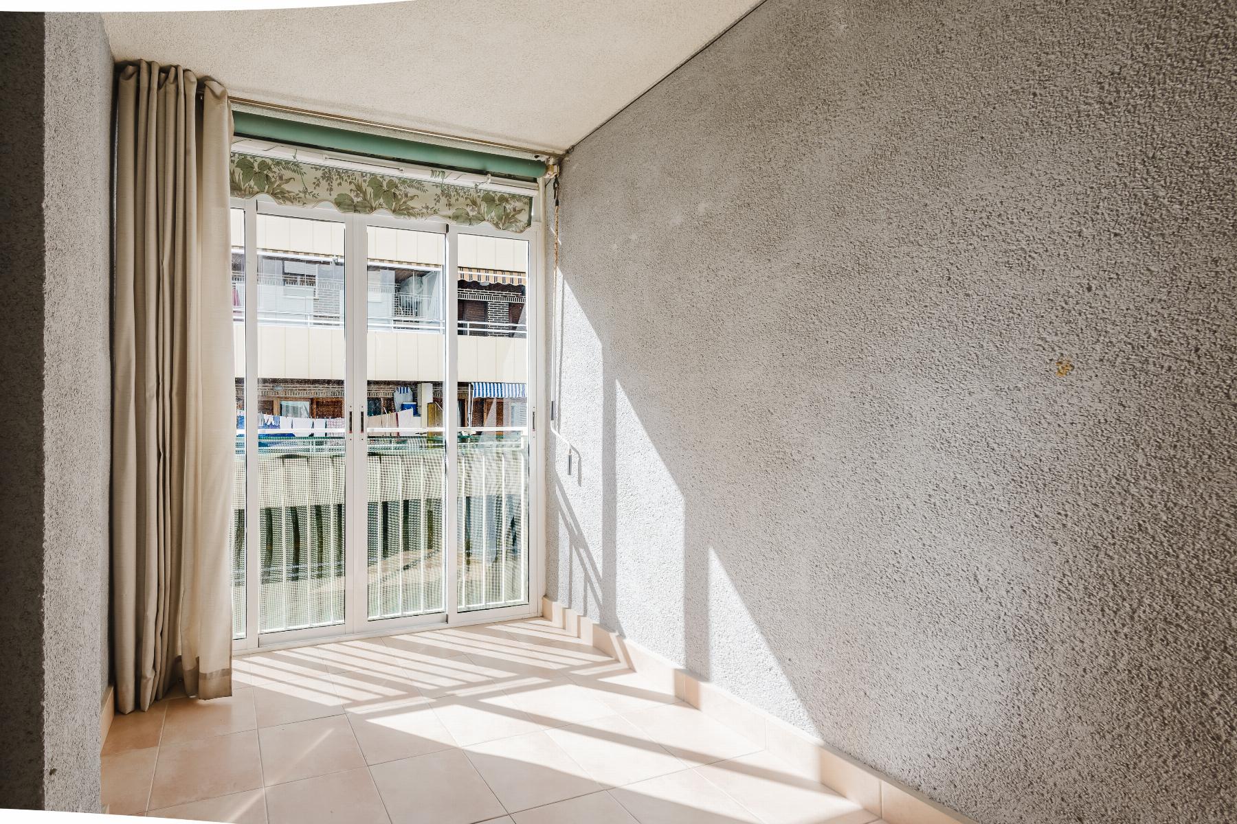 Sunlit room with beige curtains beside a sliding glass door opening to a balcony; textured gray walls and tiled floor casting striped shadows.