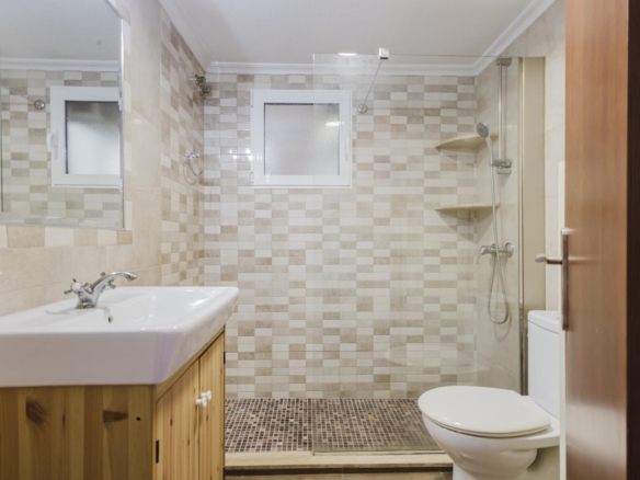 Small modern bathroom with mosaic tile walls, glass shower enclosure, white sink on a wooden vanity, and a toilet. Two-tone beige tiles and a frosted window provide natural light.