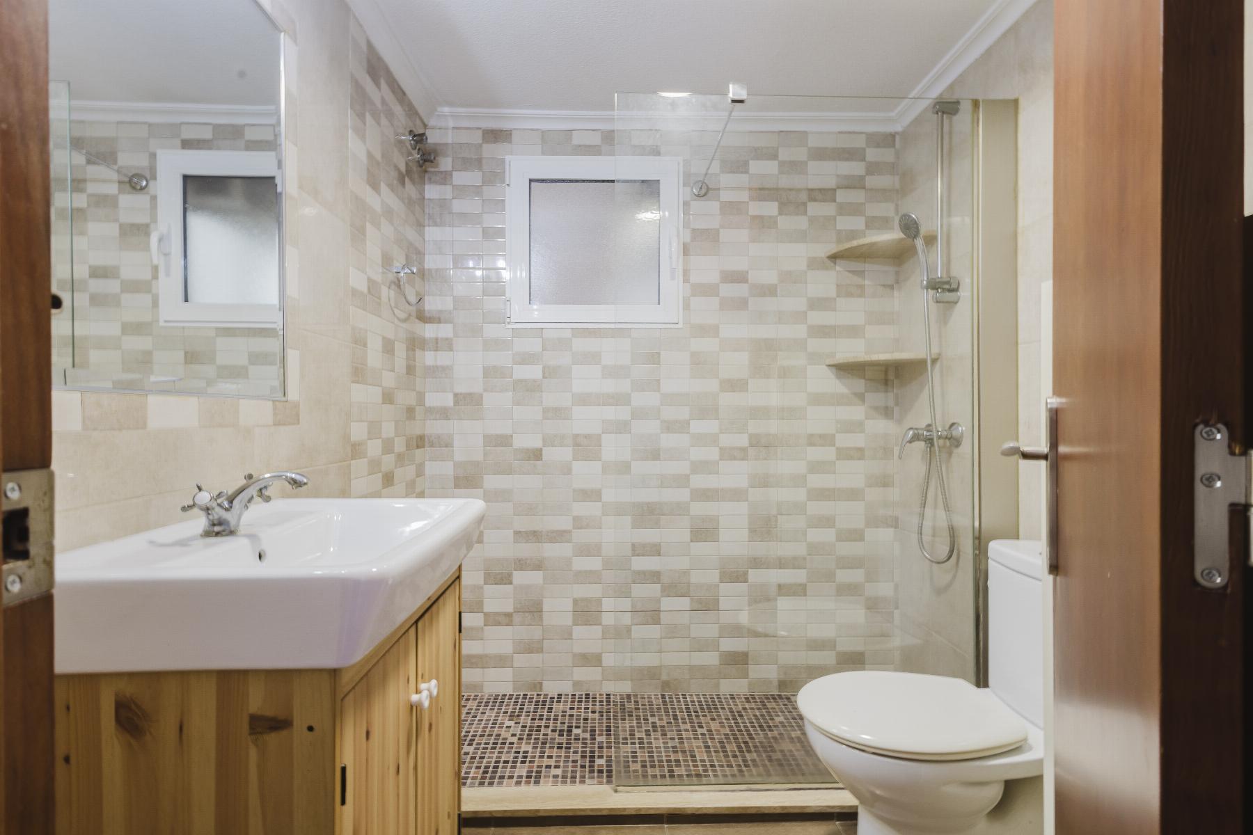 Small modern bathroom with mosaic tile walls, glass shower enclosure, white sink on a wooden vanity, and a toilet. Two-tone beige tiles and a frosted window provide natural light.