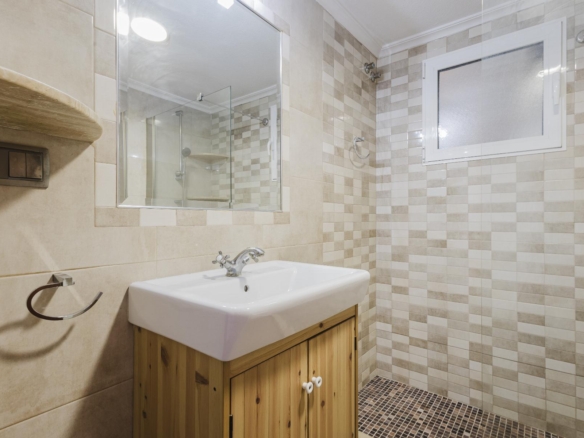 Bathroom with a white sink on a wooden vanity, a large mirror, and beige-tiled walls with a glass shower enclosure at the back.