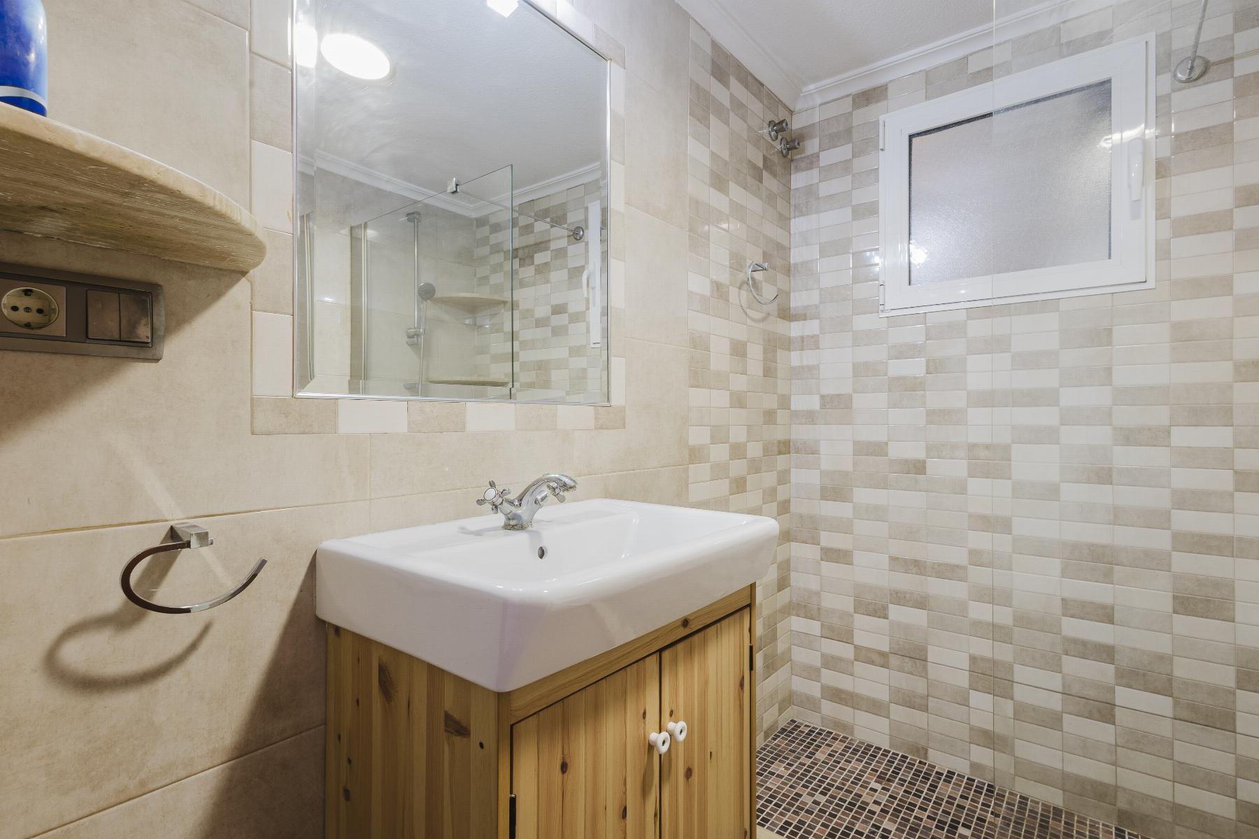 Bathroom with a white sink on a wooden vanity, a large mirror, and beige-tiled walls with a glass shower enclosure at the back.
