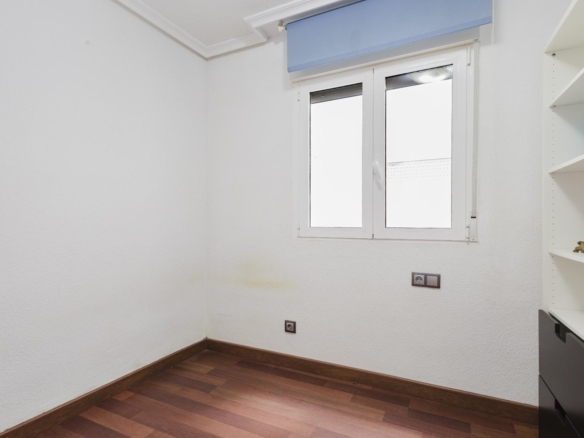 Empty small room with white textured walls, dark wood floor, and a window with a blue roller blind on the right wall; shelving unit visible on the far right.