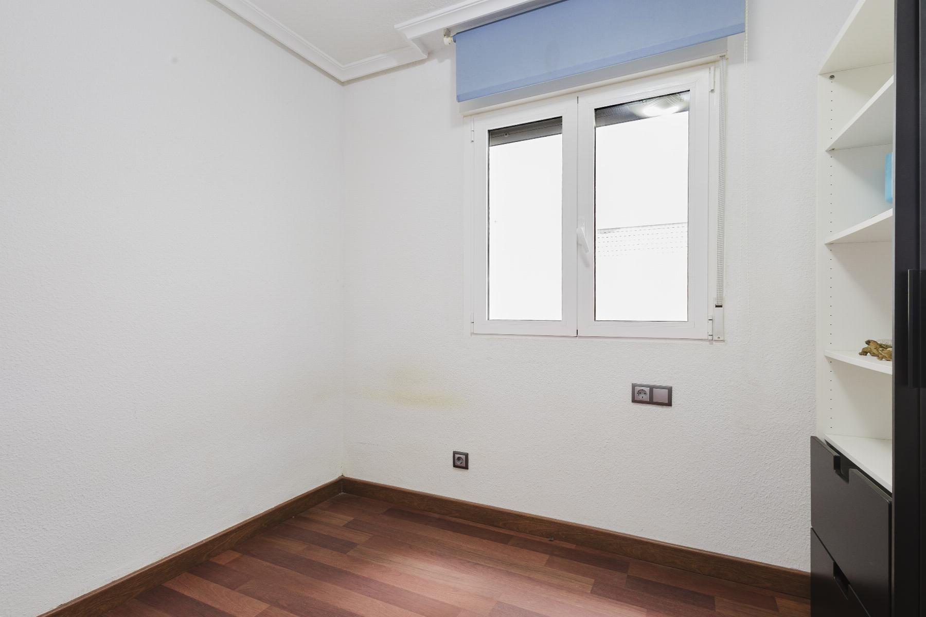 Empty small room with white textured walls, dark wood floor, and a window with a blue roller blind on the right wall; shelving unit visible on the far right.