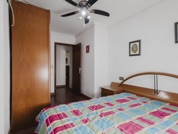 A bedroom with a striped colorful quilt on a wooden bed, a large wooden wardrobe on the left, and an open door leading to another room; ceiling fan above.
