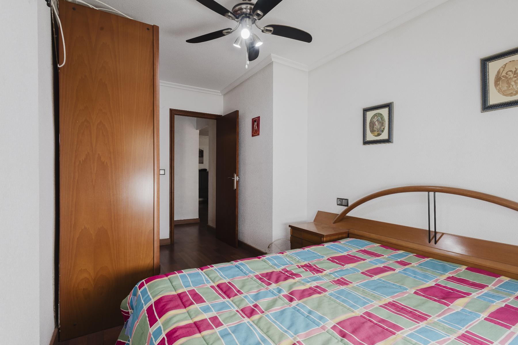 A bedroom with a striped colorful quilt on a wooden bed, a large wooden wardrobe on the left, and an open door leading to another room; ceiling fan above.