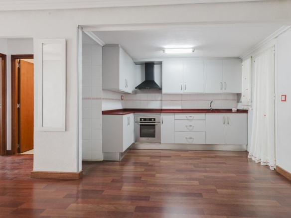 Open-plan kitchen with white cabinets, red counter, built-in oven, and black range hood against a white-tiled backsplash.