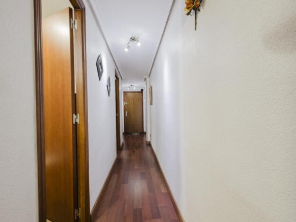 Narrow interior hallway with wooden floors, white textured walls, and multiple wooden doors along both sides, lit by ceiling spotlights.