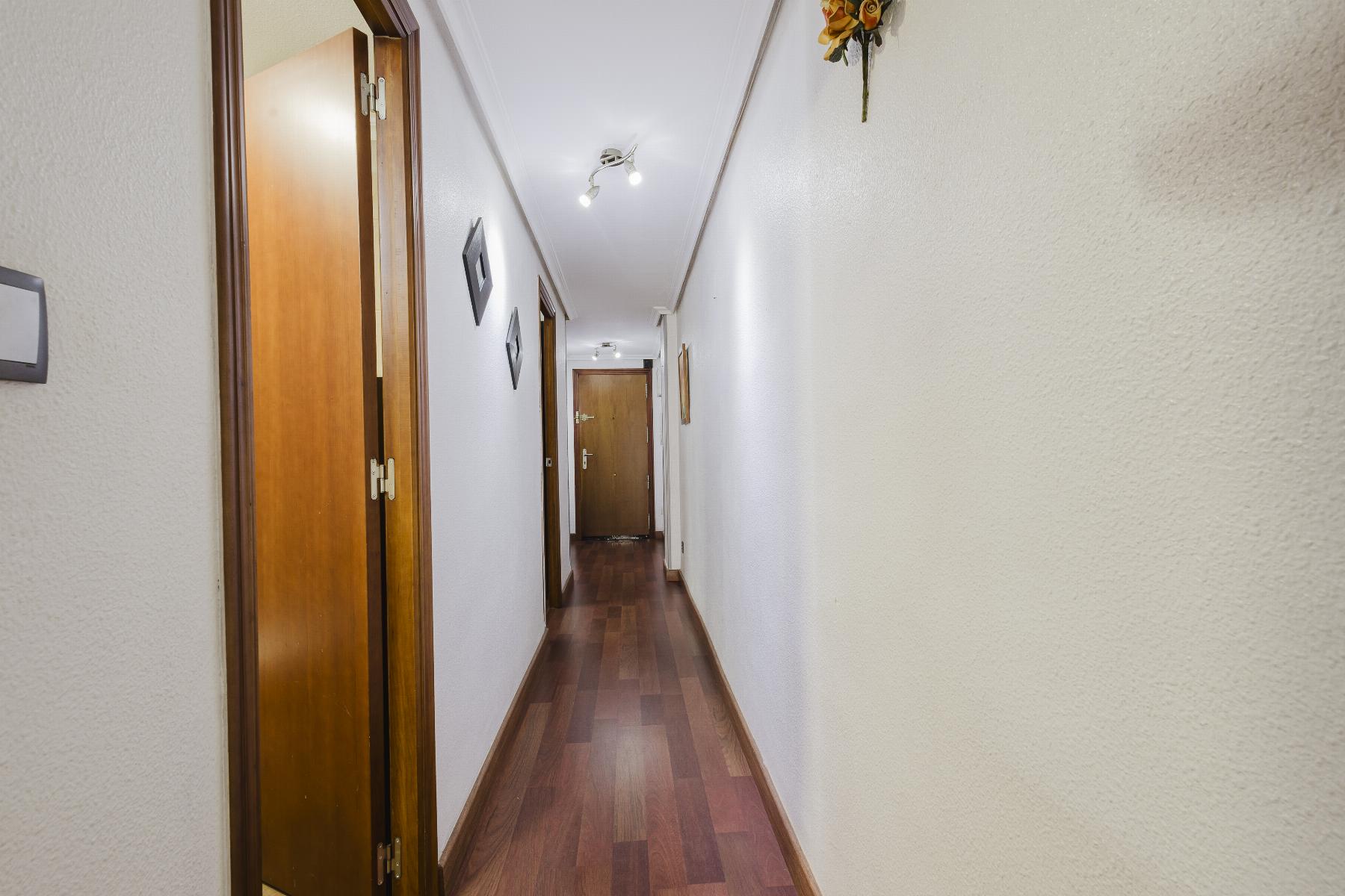 Narrow interior hallway with wooden floors, white textured walls, and multiple wooden doors along both sides, lit by ceiling spotlights.