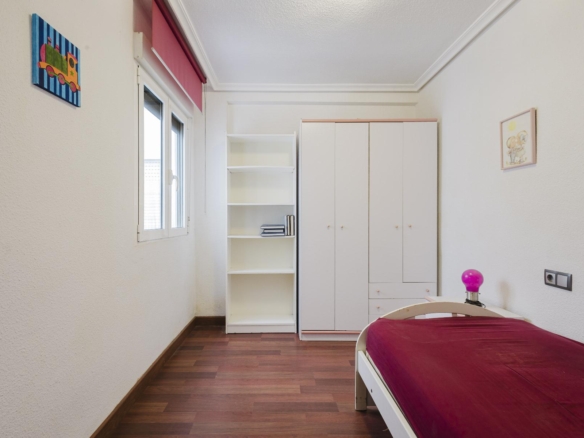 A tidy bedroom with a white wardrobe, open bookshelf, and bed with a burgundy bedspread next to a window.