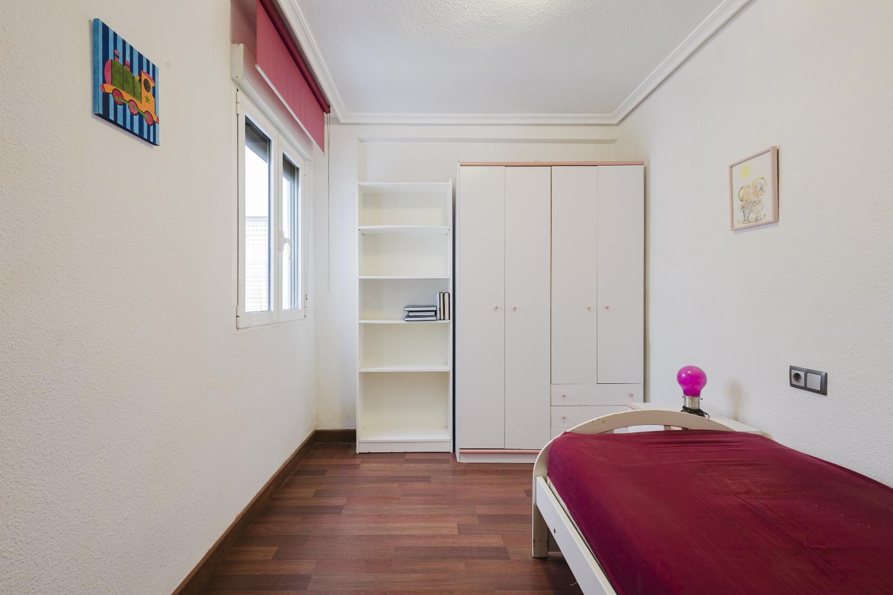 A tidy bedroom with a white wardrobe, open bookshelf, and bed with a burgundy bedspread next to a window.