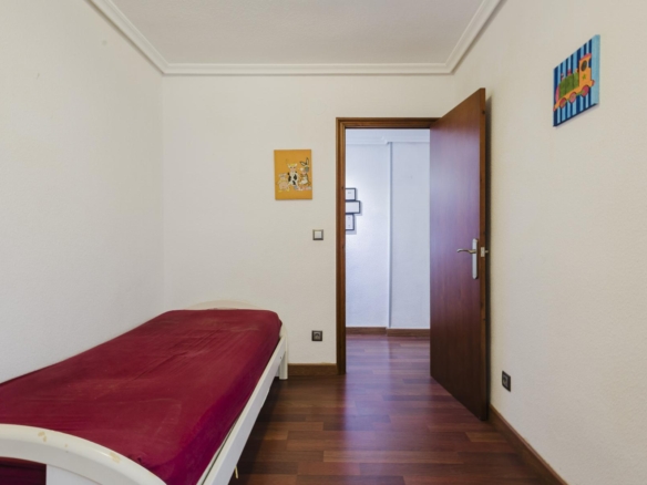 A small bedroom with a white bed covered in a red blanket along the left wall, wood floor, and an open wooden door leading to a hallway.