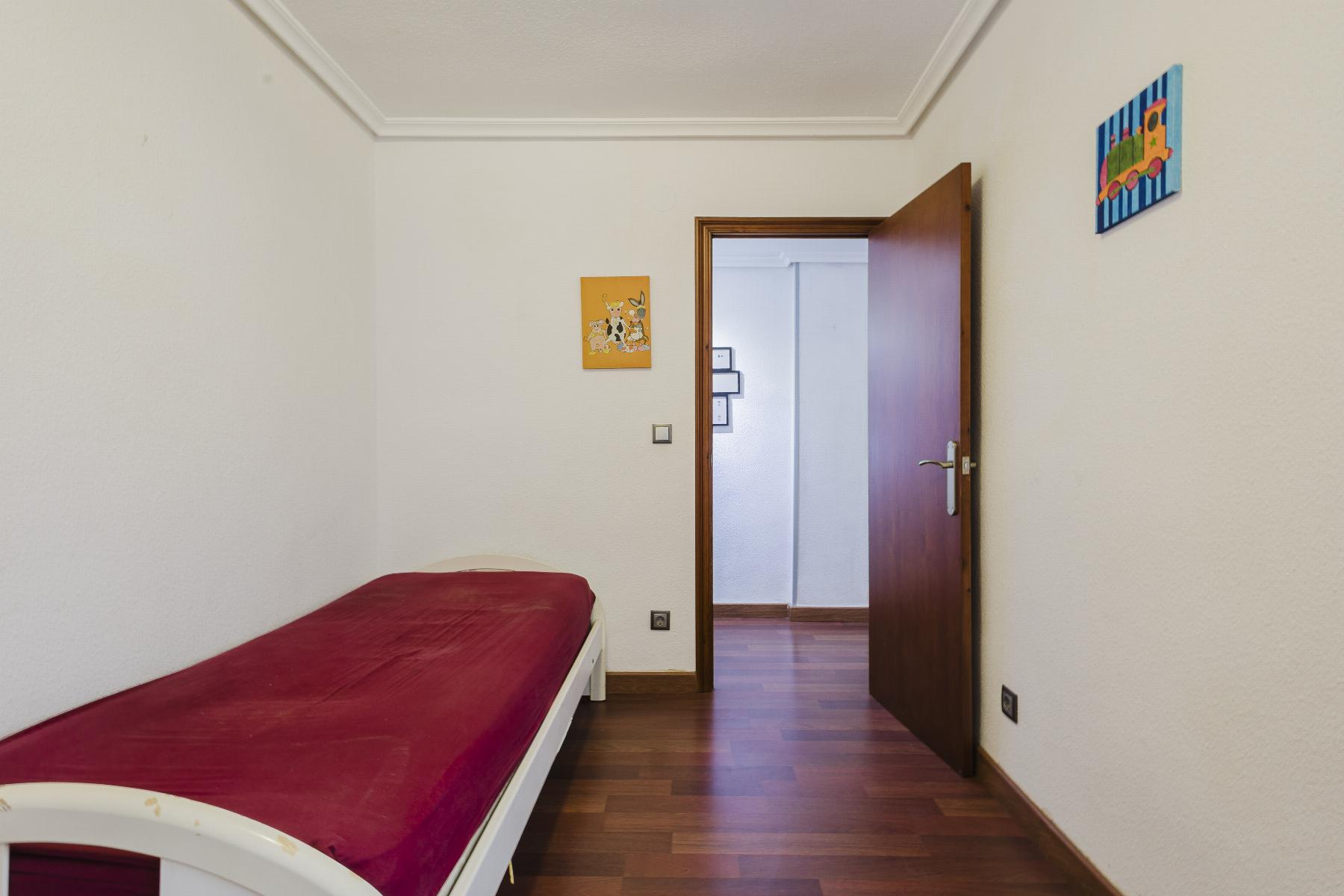 A small bedroom with a white bed covered in a red blanket along the left wall, wood floor, and an open wooden door leading to a hallway.