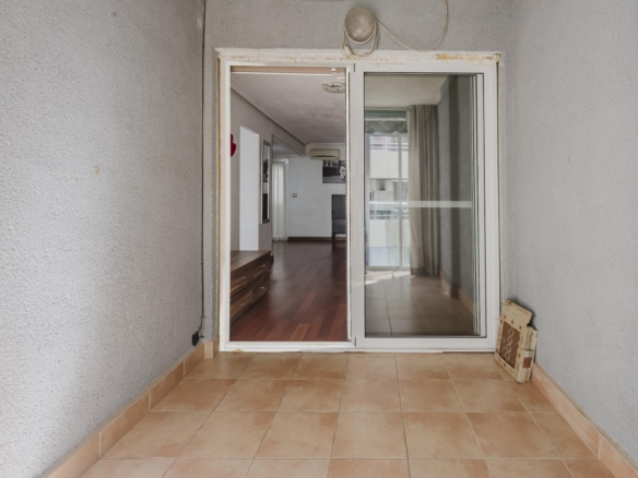 Tiled balcony area opening into a living room through a large glass sliding door; interior shows hardwood flooring and curtains.