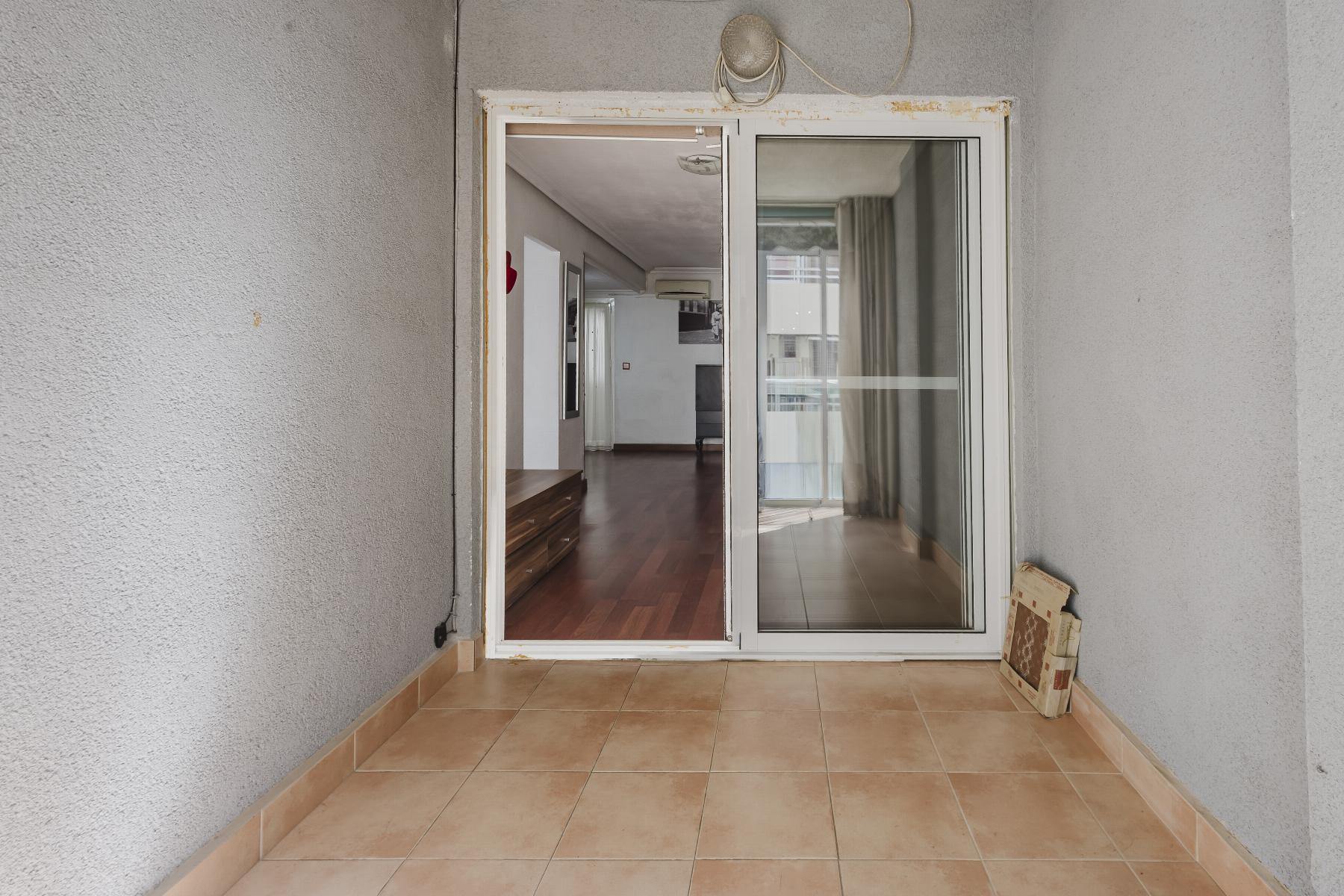 Tiled balcony area opening into a living room through a large glass sliding door; interior shows hardwood flooring and curtains.