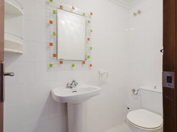 White tiled bathroom with a pedestal sink and a square mosaic-framed mirror bordered by orange and green tiles above the sink.