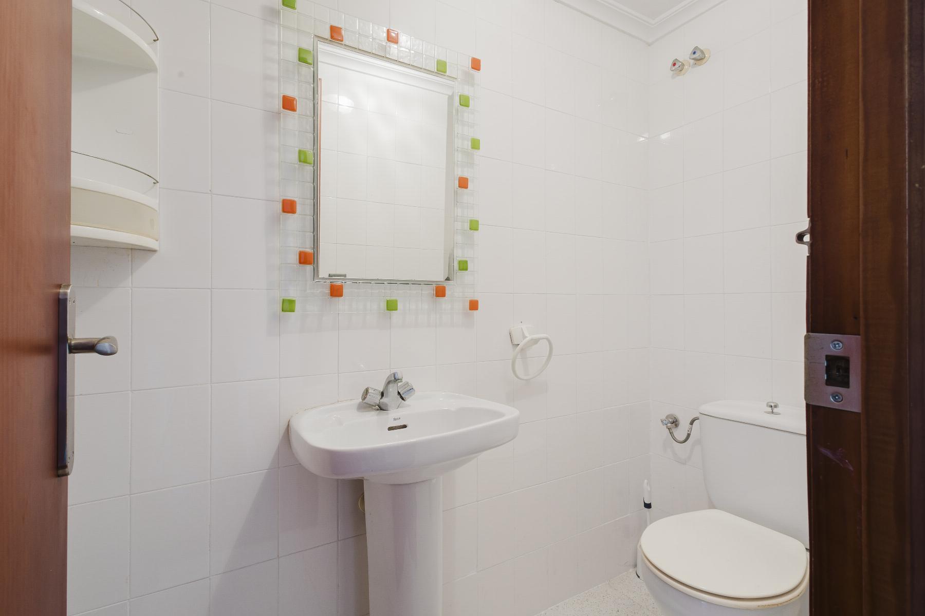 White tiled bathroom with a pedestal sink and a square mosaic-framed mirror bordered by orange and green tiles above the sink.