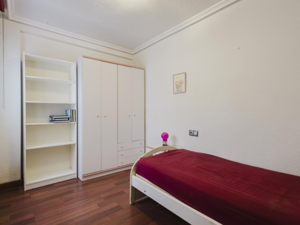 A white wardrobe and an open white bookshelf along a wall of a tidy bedroom, with a bed on the right covered in a burgundy blanket and a pink bedside lamp nearby.