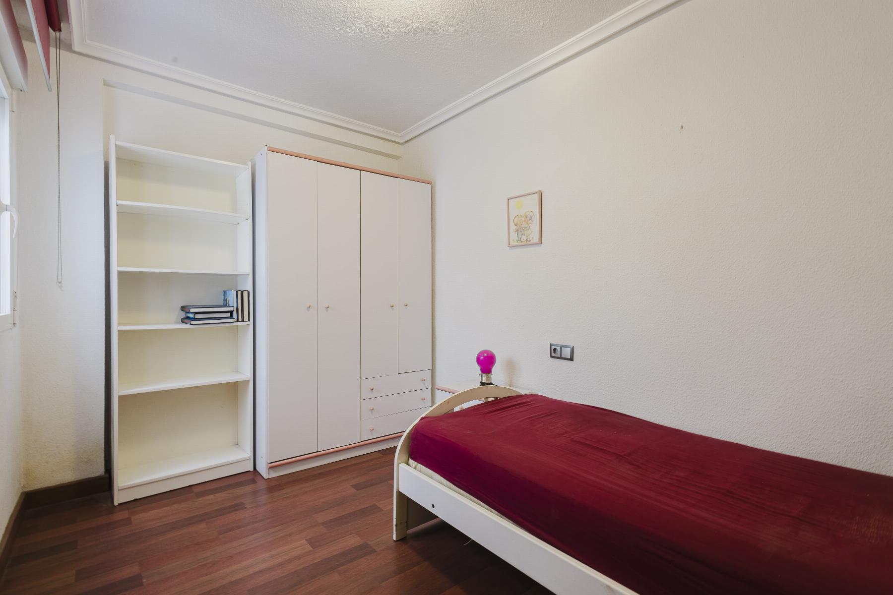 A white wardrobe and an open white bookshelf along a wall of a tidy bedroom, with a bed on the right covered in a burgundy blanket and a pink bedside lamp nearby.