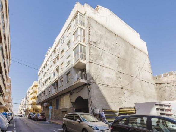 Narrow street in a city with parked cars and mid‑rise buildings; a tall white building with balconies dominates the right side under a clear blue sky.