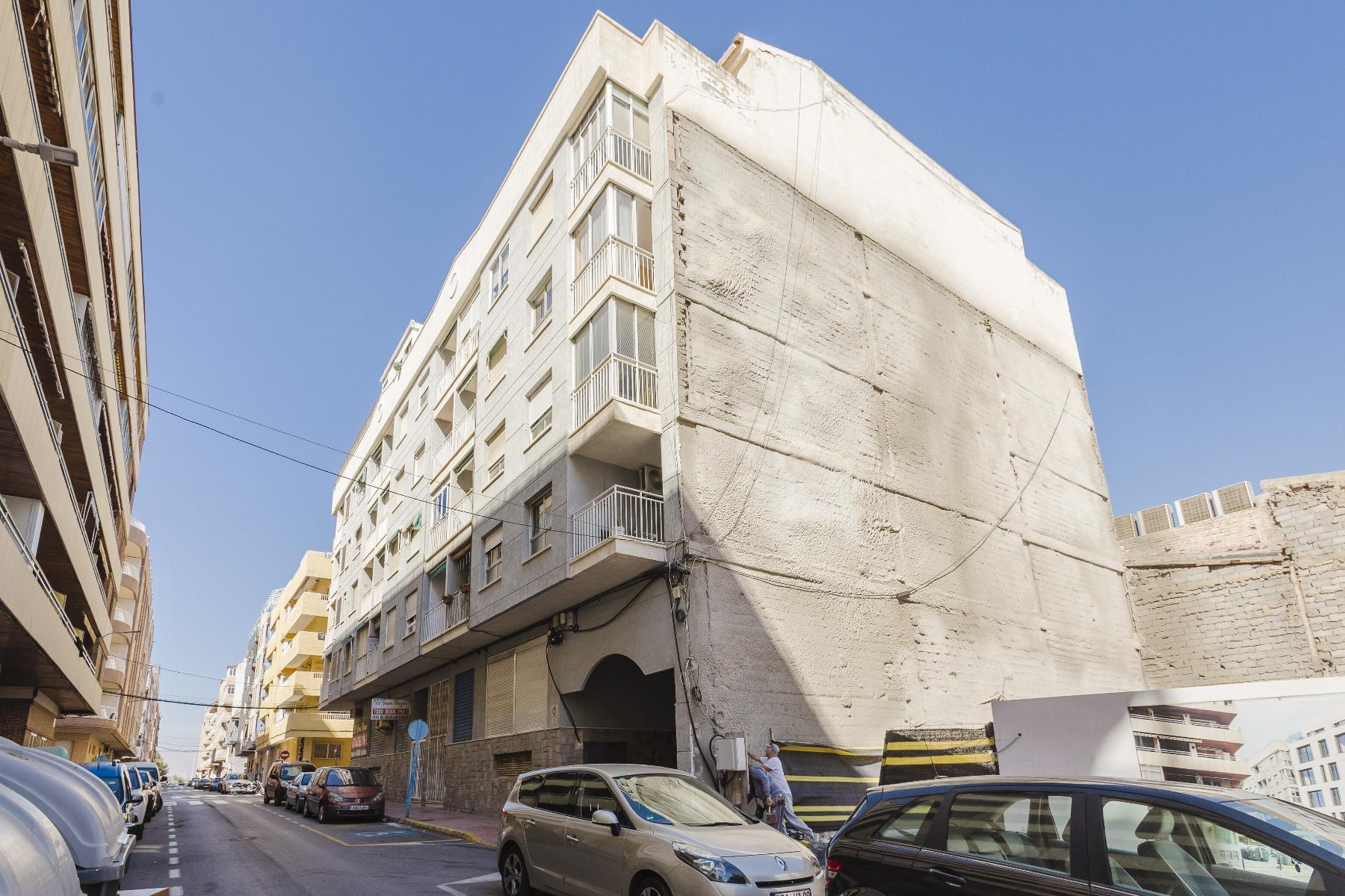 Narrow street in a city with parked cars and mid‑rise buildings; a tall white building with balconies dominates the right side under a clear blue sky.