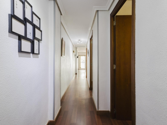 Long hallway with white textured walls, framed pictures on the left, wooden doors on the right, and a bright doorway at the far end.