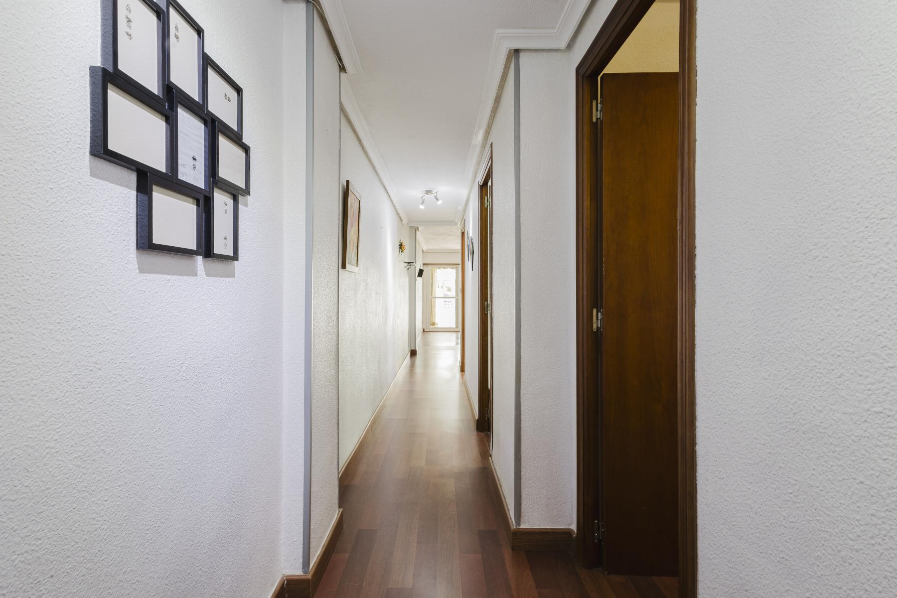 Long hallway with white textured walls, framed pictures on the left, wooden doors on the right, and a bright doorway at the far end.