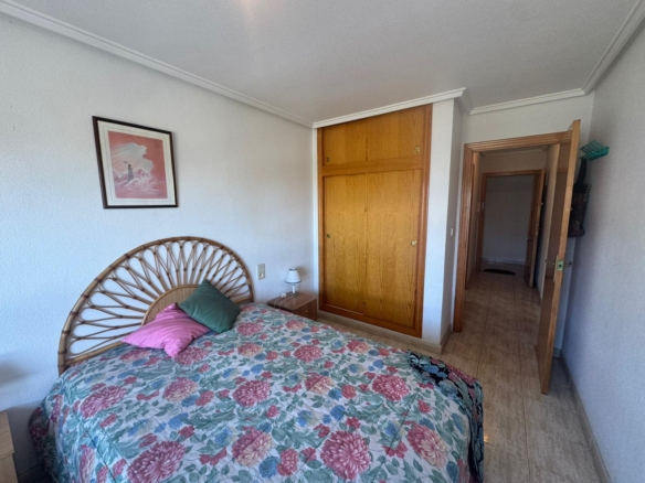 Bedroom with a floral quilt on a wicker bed, wooden built-in wardrobe, small nightstand and lamp, and a framed pink artwork on the wall.