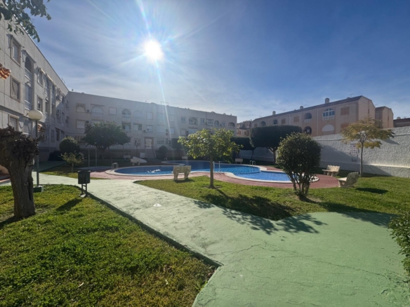 Sunlit residential courtyard with a blue kidney-shaped pool, green grass, and surrounding apartment buildings.