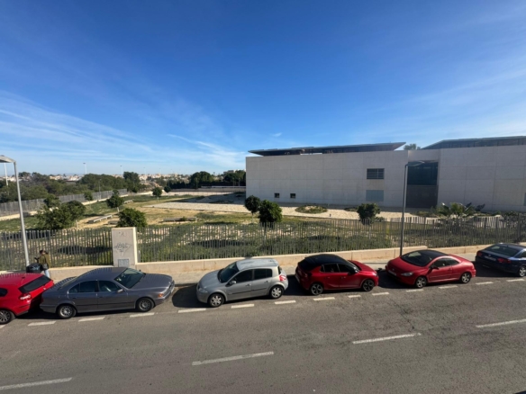 Street view with parked cars in front of a modern white building and a grassy open area under a clear blue sky