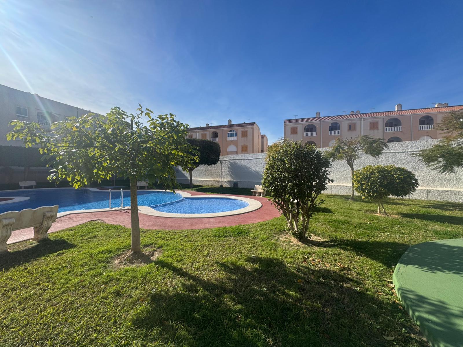 Sunlit hotel courtyard with a curved blue pool, green grass, and trimmed trees, behind beige apartment buildings with red-tiled roofs.