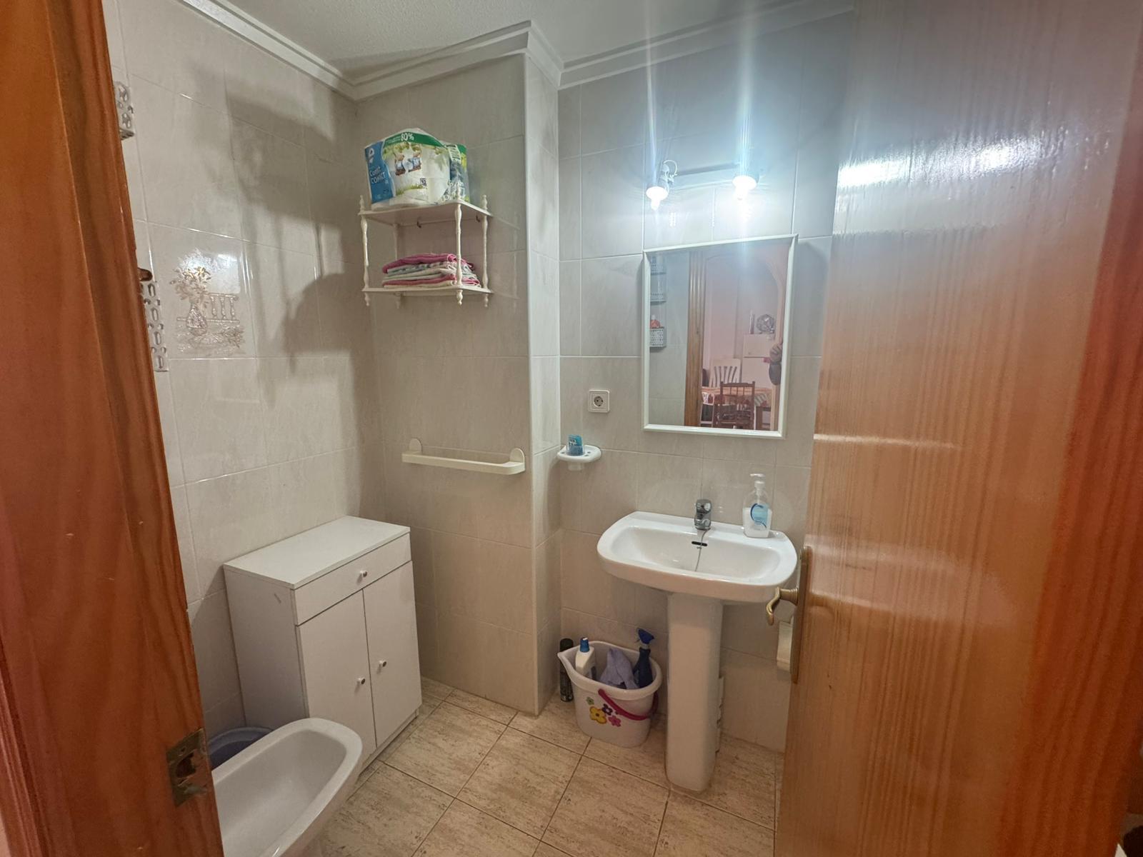 Small beige-tiled bathroom with a white sink, mirror, toilet, wall shelf with towels, and a cleaning bucket on the floor.