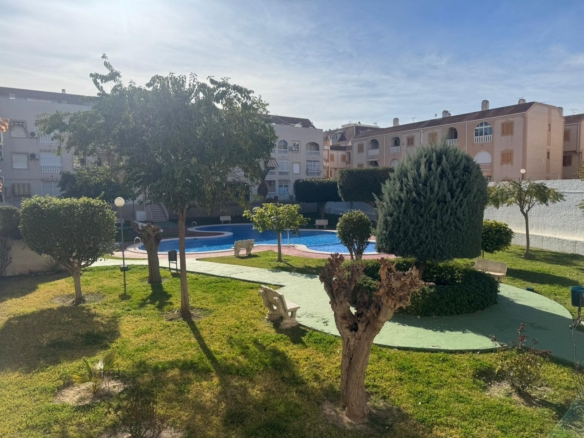 Residential courtyard with a curved blue pool, manicured bushes, and benches, surrounded by beige apartment buildings under a clear sky.