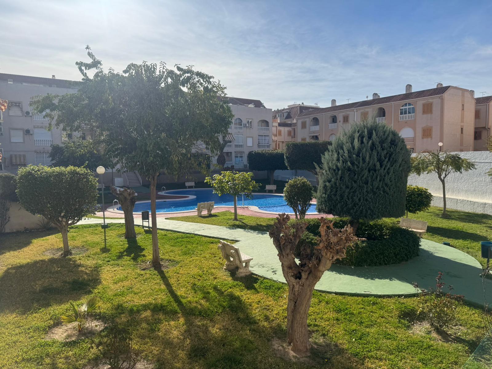 Residential courtyard with a curved blue pool, manicured bushes, and benches, surrounded by beige apartment buildings under a clear sky.