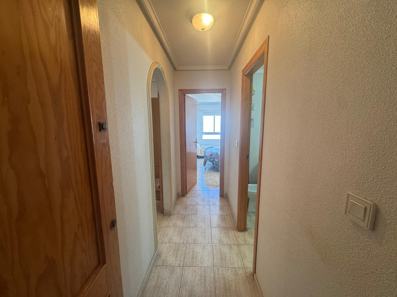 Narrow hallway with wooden doors on both sides, tiled floor, and a bedroom visible at the end under a ceiling light